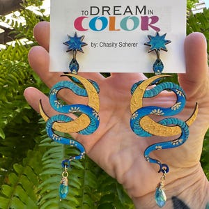 May include: A pair of long, blue and gold snake earrings with a star-shaped top and a teardrop-shaped crystal charm. The earrings are held in front of a sign that says "TO DREAM IN COLOR" by Chasty Scherer.