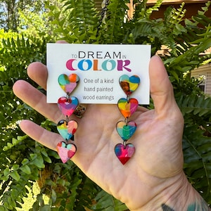 May include: A pair of colorful wooden heart earrings with a layered design. The earrings are hand painted and feature a variety of bright colors, including red, orange, yellow, green, blue, and purple. The earrings are labeled "To Dream in Color" and "One of a kind hand painted wood earrings".