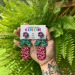 May include: A pair of pink and turquoise geometric earrings with a leopard print design. The earrings are made of clay, resin, and wood. The earrings are on a card that says "To Dream in Color".