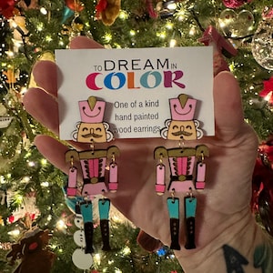 May include: A pair of hand-painted wooden nutcracker earrings. The earrings are pink, blue, and gold with a whimsical design. The earrings are labeled "TO DREAM IN COLOR" and "One of a kind hand painted wood earrings".