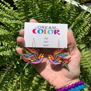May include: Rainbow colored wooden bird earrings with gold hoops. The earrings are on a card that says "To Dream in Color" with the words "clay, resin, wood" listed below.
