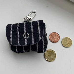 Può includere: Una piccola custodia nera con strisce verticali bianche, dotata di chiusura a scatto in metallo argentato e moschettone argentato. La custodia è mostrata accanto a tre monete in euro, nello specifico da 5, 10 e 20 centesimi.