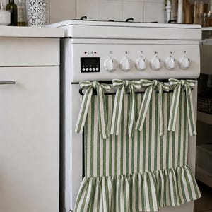 Pode incluir: Um fogão branco com uma capa de tecido às riscas verdes e brancas colocada sobre a porta do forno. A capa é fixada com quatro laços de tecido atados no topo e apresenta uma bainha com folhos, acrescentando um elemento têxtil decorativo.