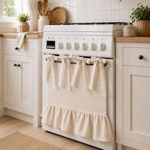 May include: A cream-coloured fabric oven cover featuring a ruffled bottom edge and four decorative tie-top bows. The textile accessory is draped over the handle of a white kitchen stove, covering the front glass panel with a textured, neutral-toned cloth.
