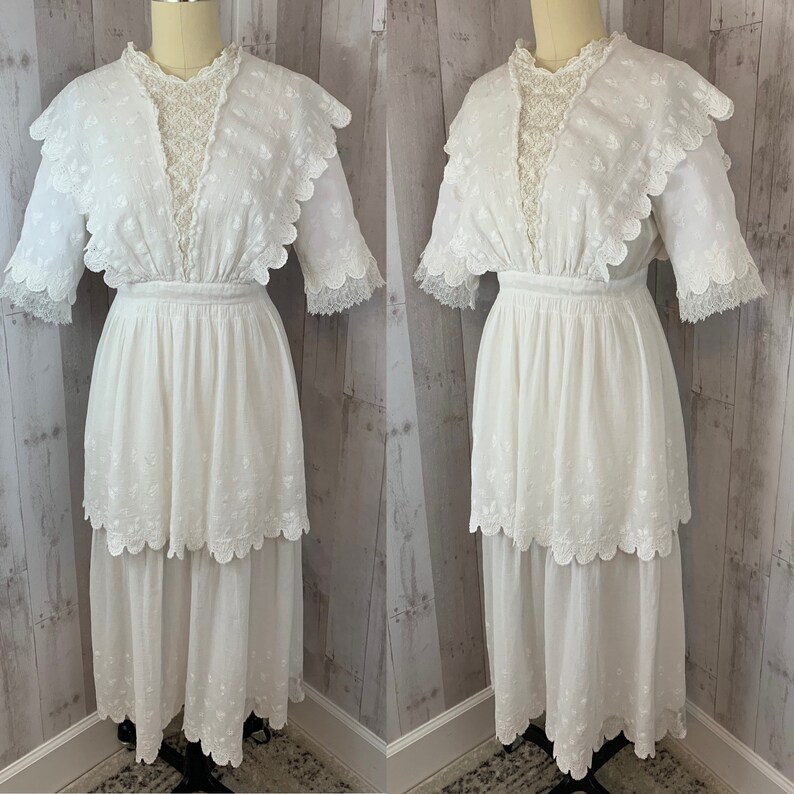 May include: A white cotton dress with a lace overlay and a tiered skirt. The dress has a scalloped neckline and short sleeves.