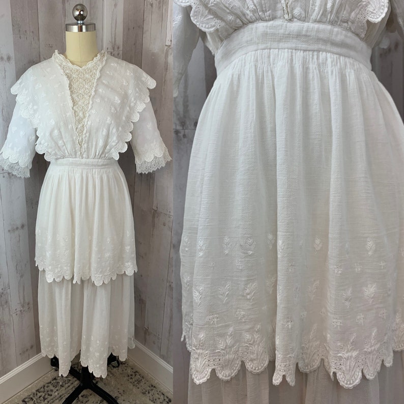 May include: A white cotton dress with a lace overlay and tiered skirt. The dress has a scalloped hem and embroidered floral details.