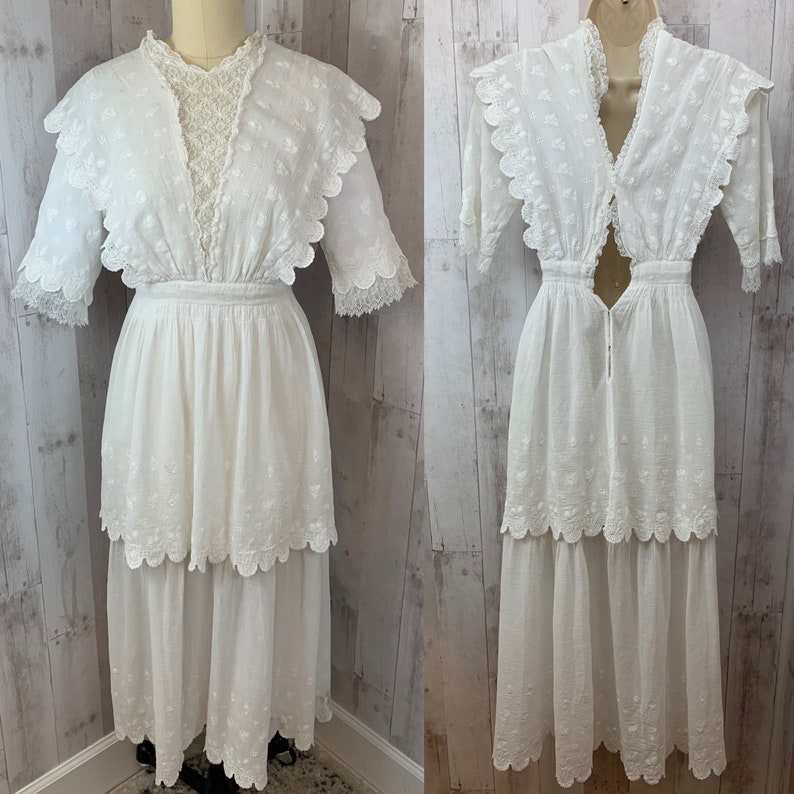 May include: A white cotton dress with a lace overlay and scalloped edges. The dress has a fitted bodice and a flowing skirt with multiple tiers. The dress has a keyhole opening at the back.