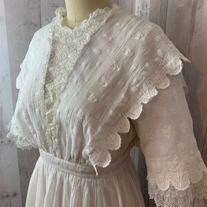 May include: A white cotton dress with a lace overlay and scalloped edges. The dress has a fitted bodice and a full skirt.