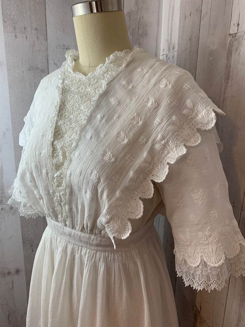 May include: A white cotton dress with a lace overlay and scalloped edges. The dress has a fitted bodice and a full skirt.