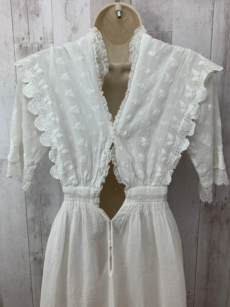 May include: A white cotton dress with a lace trim and a keyhole back. The dress has a v-neck and a fitted bodice. The skirt is full and flowing.