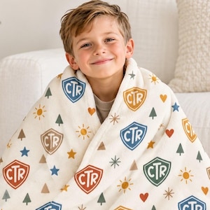 May include: A cream-colored plush blanket featuring a repeating pattern of shield-shaped emblems with the letters CTR inside. The design includes small icons of suns, hearts, stars, triangles, and pine trees in blue, orange, and green.