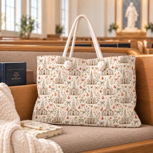 May include: A tote bag featuring a repeating pattern of tan church buildings, small orange leaves, and green bows. The bag has two thick, white twisted rope handles attached with white pom-poms at the base of each handle on the front panel.