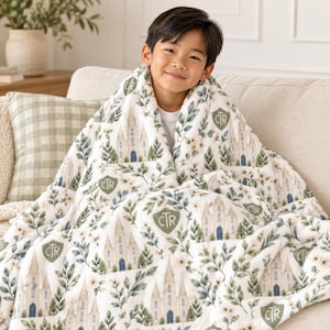 May include: A white plush blanket featuring a repeating pattern of beige buildings with blue doors, green leafy branches, small flowers, and olive green shield-shaped emblems containing the letters CTR in a dark green font.