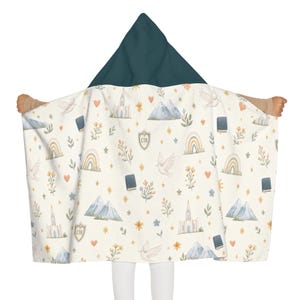May include: A hooded blanket with a dark teal hood and a cream-colored body featuring a repeating pattern of white doves, blue mountains, rainbows, small books, floral sprigs, orange hearts, yellow stars, and shields with the letters CTR printed on them.