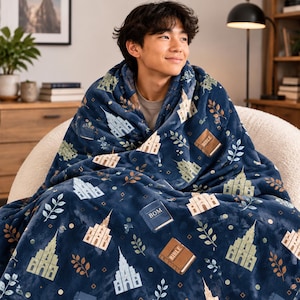 May include: A plush navy blue throw blanket featuring a repeating pattern of light-colored temple illustrations, brown books labeled BIBLE, and blue books labeled BOM, interspersed with small green leaf sprigs and geometric diamond shapes.