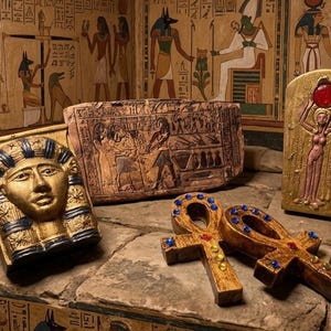 May include: A collection of Egyptian-themed decorative items including a gold and blue pharaoh mask, a rectangular stone tablet with hieroglyphics, a gold-toned plaque featuring a figure with a red sun disk, and two wooden ankh symbols with blue gems.