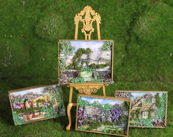 May include: Miniature framed landscape art featuring quaint cottages with thatched roofs and lush gardens. The artwork is displayed on a gold easel and includes three additional framed pieces, all set against a backdrop of green moss.