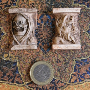 May include: Two small, rectangular, off-white sculptures with carved faces, one a skull and the other a bearded figure. A Euro coin is next to the sculptures. The background is a patterned design in gold, blue, and brown.