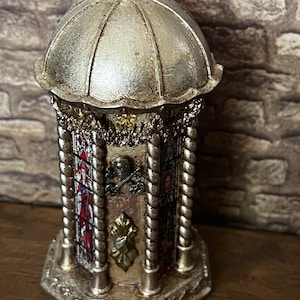 Op de afbeelding: Een decoratieve zilveren en gouden toren met een koepelvormige top en glas-in-loodramen. De toren heeft een gedetailleerd ontwerp met ingewikkelde houtsnijwerk en een touwachtig patroon.