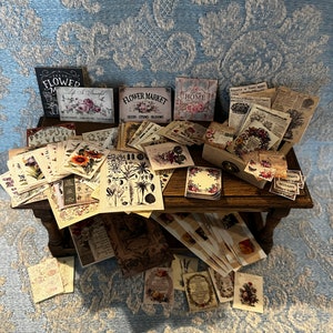 May include: A wooden table piled high with vintage-style paper ephemera, including cards, tags, and labels. The papers feature floral designs, botanical illustrations, and text such as "Flower Market" and "Home".
