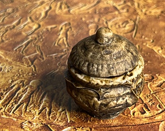 May include: A small, lidded jar with a textured, aged appearance. The jar is primarily a muted brown with lighter, cream-colored accents. The lid features a small, rounded knob. The background has a carved relief pattern.