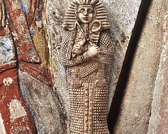 May include: A beige ceramic sculpture of an Egyptian pharaoh wearing a headdress and holding a staff. The sculpture is detailed with hieroglyphics and has a textured surface.