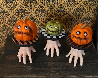 Könnte beinhalten: Drei Halloween-Dekorationen in Form von geschnitzten Kürbissen. Jeder Kürbis mit einem geschnitzten Gesicht sitzt auf einer kleinen Plattform, die von einer handförmigen Basis getragen wird. Die Kürbisse sind orange und grün, mit schwarzen und weißen Details.
