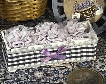 May include: A small, rectangular box with a black and white checkered pattern, trimmed with white lace and a purple bow. The box contains lavender-colored lace and sits on a patterned tile surface. A basket of purple yarn and a decorative wheel are in the background.