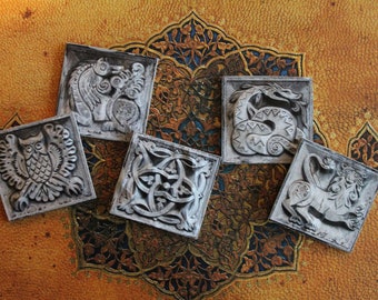 May include: Five square ceramic tiles with raised, detailed carvings of mythical creatures. The tiles are white and have a textured surface. The tiles are arranged in a circle on a gold and blue patterned surface.