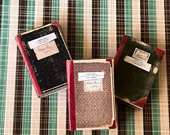 May include: Three vintage hardcover journals with brown, black, and red covers. The journals have a checkered pattern and are on a green and brown plaid surface.
