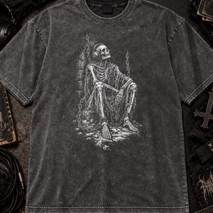 May include: A charcoal grey acid-wash t-shirt featuring a white graphic print of a skeleton wearing headphones, sitting in a chained, stone-walled environment. The shirt has a crew neckline, short sleeves, and a distressed, vintage-inspired texture.