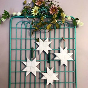 Puede incluir: Cuatro estrellas de papel blanco cuelgan de una rejilla metálica turquesa. La rejilla está decorada con una guirnalda floral de flores blancas, rosas y amarillas.