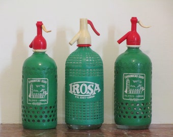 May include: Three green glass soda siphon bottles with red and white plastic tops. The bottles have a grid pattern and are labelled with the text "Carbonicas Guiu", "Tel 211572 - Lerida", "Irosa", and "FTE 3345 - Lerida".