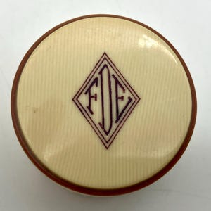 Puede incluir: Un estuche redondo de color crema con una textura fina de rayas verticales. En el centro de la tapa hay un emblema en forma de diamante de color morado oscuro que contiene el monograma FDE en letras serif negritas.