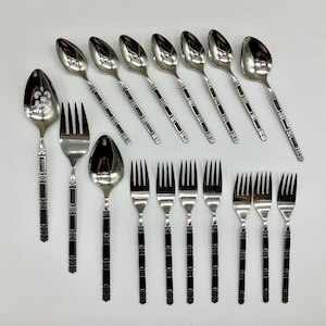 May include: A set of stainless steel flatware consisting of seven spoons and seven forks. Each piece features a polished silver finish with a decorative handle design containing black rectangular inlays and small floral accents.