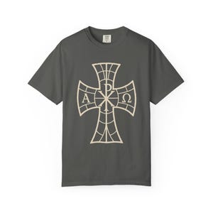 Puede incluir: Una camiseta de manga corta gris carbón que presenta un gráfico grande de color crema de una cruz con un patrón de cuadrícula. El centro de la cruz muestra el símbolo Chi-Rho, flanqueado por las letras griegas Alfa y Omega a los lados.