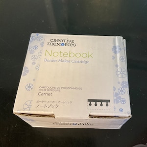 Puede incluir: Caja blanca con un patrón de flores azules. La caja contiene un cartucho de borde de cuaderno Creative Memories. El texto en la caja dice "Notebook Border Maker Cartridge" y "Carnet".