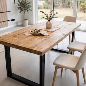 Puede incluir: Una mesa de comedor rectangular con tablero de madera natural y patas gruesas de metal negro en forma de U. La mesa está dispuesta con dos servilletas de tela beige, un pequeño jarrón de cerámica con ramas de eucalipto y tres lámparas colgantes.