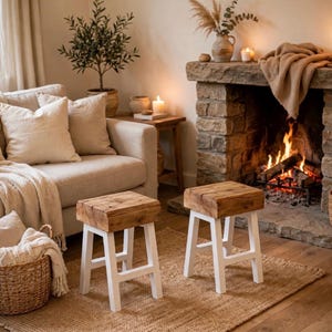 Puede incluir: Dos taburetes pequeños con asientos gruesos de madera en tono natural con vetas y texturas visibles, sostenidos por cuatro patas de madera resistentes pintadas de blanco. Los taburetes tienen un diseño simple y funcional con base cuadrada.