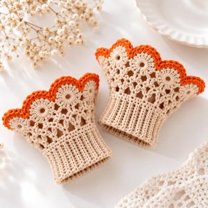 Puede incluir: Un par de muñequeras de ganchillo en color beige con una base acanalada, un intrincado patrón de encaje calado y un borde superior festoneado acabado con un ribete de hilo naranja vibrante. El diseño incluye motivos circulares en el encaje.