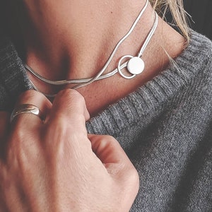 Puede incluir: Un collar de cadena de plata con un colgante redondo. El collar está en capas y tiene un diseño delicado.