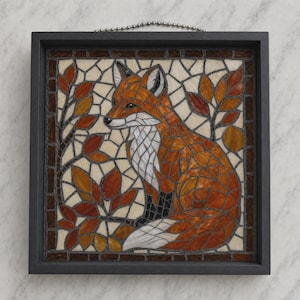 May include: A square wall hanging featuring a mosaic of a fox surrounded by autumn leaves. The fox is rendered in shades of orange, white, and black glass pieces. The background consists of cream-colored tiles, and the entire mosaic is encased in a dark gray frame with a metal ball chain for hanging.