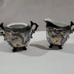 May include: A ceramic sugar bowl and creamer set featuring a black and white gradient finish with raised dragon motifs. Both pieces are supported by three small feet and include ornate handles, with the creamer featuring a pouring spout.