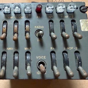 Puede incluir: Un panel de control de radio de avión gris con múltiples interruptores de palanca y botones iluminados. El panel incluye etiquetas como VHF, PA, INT, HF, NAV, MKR, ADF y VOICE. Tiene una carcasa metálica rectangular con tornillos visibles en las esquinas.
