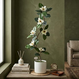 May include: A tall artificial magnolia tree with dark green leaves and five white blossoms, planted in a simple white cylindrical ceramic pot. The plant stands on a woven circular mat next to a stack of books and a small analogue clock.