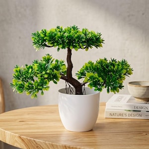 Puede incluir: Un bonsái artificial con follaje verde vibrante y un tronco marrón oscuro, plantado en una maceta de cerámica blanca lisa. La planta presenta ramas en capas con hojas densas en forma de aguja, creando una apariencia estructurada.