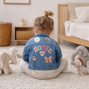 Puede incluir: Una chaqueta de mezclilla azul claro con parches bordados coloridos en la espalda, que incluyen una margarita blanca, un arcoíris, el nombre DOVIC en letras pastel, un corazón con signo de paz, una cara sonriente y una mariposa rosa y naranja.