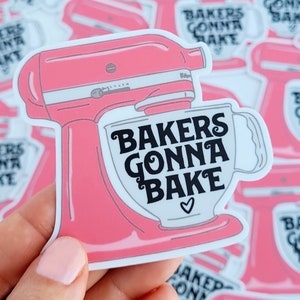 May include: A pink sticker featuring a stand mixer design with the text "Bakers Gonna Bake" in black script. The sticker has a white border and a small heart symbol. The image is of a sticker held by a person.