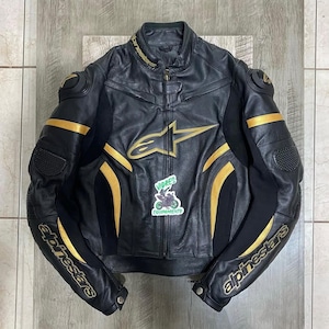Può includere: Una giacca da moto in pelle nera con accenti dorati e il logo Alpinestars sul petto, sul colletto e sulle maniche. La giacca presenta una cerniera centrale, imbottiture protettive su spalle e gomiti e un adesivo con il testo VIDRES EQUIPAMIENTO.