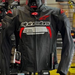 May include: A black leather motorcycle jacket with red and white accents. The chest features the Alpinestars logo and the text ONE GOAL. ONE VISION. It includes shoulder armour, a mandarin collar, and a full-length front zip with hanging tags.